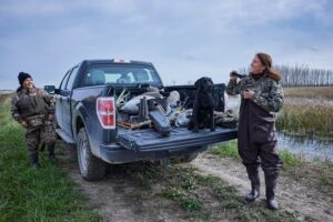 Two women on either side of a mud-covered black truck with gear and a black dog in tow prepare for a day of duck hunting.