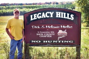 Photo of Dick Mielke at the sign on the edge of his property christening it 'Legacy Hills.'