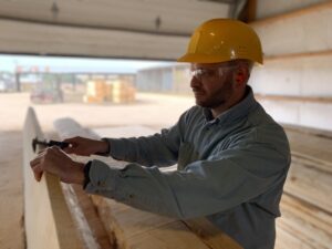 A man wearing a yellow hard hat and safety glasses measures a piece of wood in a lumberyard with stacks of wood in the background.