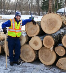 A man in a reflective vest stands next to a large stack of tree logs in a snow-covered yard.