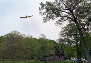 A wide view showing an airplane spraying pesticide over Devils Lake State Park to control spongy moth caterpillars.